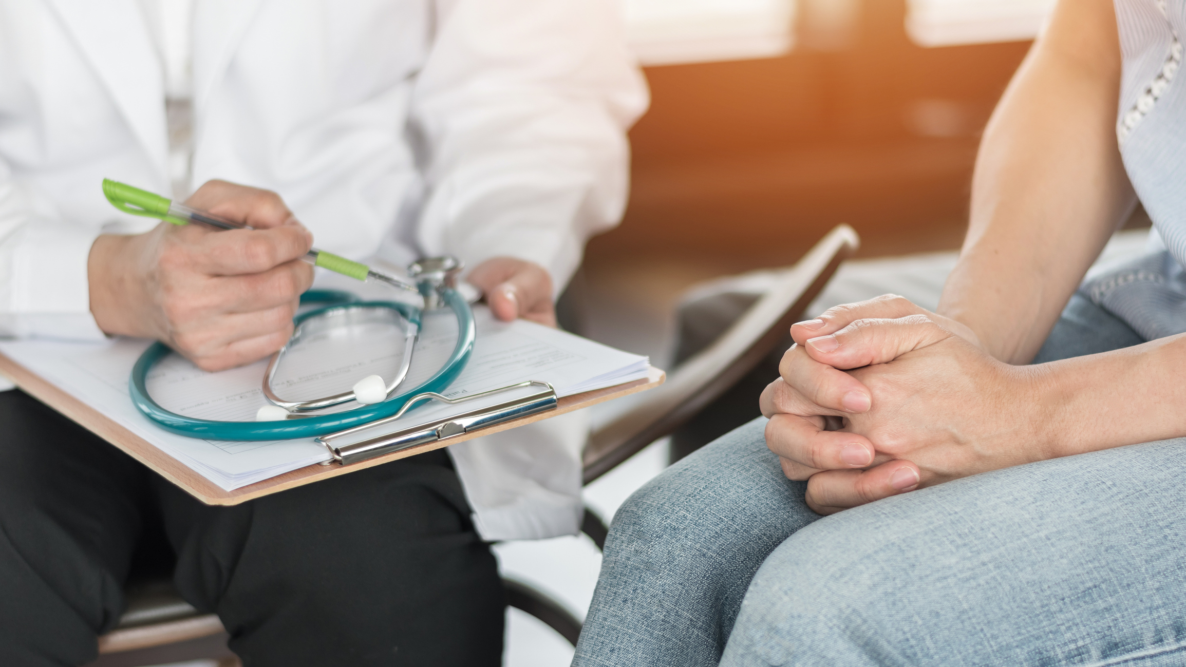 Picture of two people's hands. One is holding a clipboard with a stethoscope sitting on it. The other appears to be a patient with their hands clasped in their lap